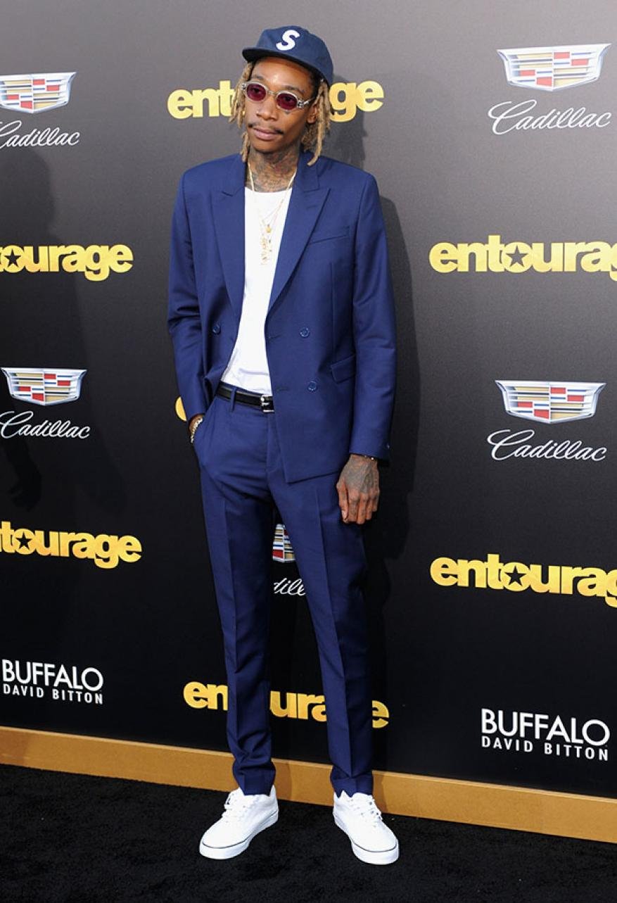 Adult male in a blue suit posing on a red carpet in front of an "Entourage" sign. He's wearing a baseball cap, white sneakers, and jewelry.