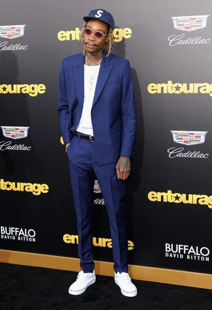 Adult male in a blue suit posing on a red carpet in front of an "Entourage" sign. He's wearing a baseball cap, white sneakers, and jewelry.
