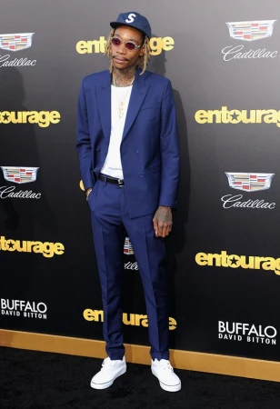 Adult male in a blue suit posing on a red carpet in front of an "Entourage" sign. He's wearing a baseball cap, white sneakers, and jewelry.