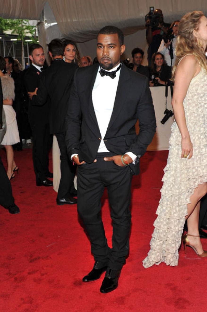 Kanye West on a red carpet wearing a black tuxedo with a white shirt and black bow tie and black shoes attends a public event.