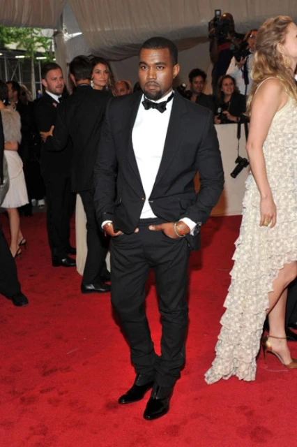 Kanye West on a red carpet wearing a black tuxedo with a white shirt and black bow tie and black shoes attends a public event.