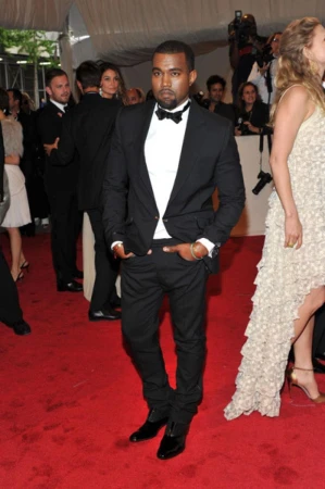 Kanye West on a red carpet wearing a black tuxedo with a white shirt and black bow tie and black shoes attends a public event.