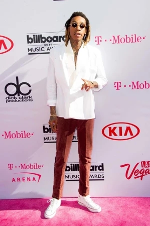 Wiz Khalifa posing at the 2016 Billboard Music Awards on pink carpet wearing a white jacket and maroon pants. Backdrop shows sponsors.