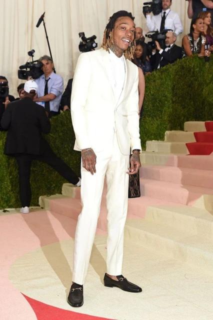 Wiz Khalifa poses in an off-white suit on a red-carpeted staircase at a celebrity event, surrounded by photographers and onlookers.
