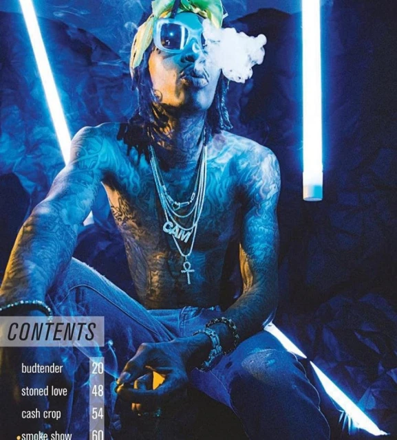 Tattooed adult male seated blowing smoke, wearing sunglasses and jewelry in a blue-lit indoor setting with magazine graphics visible.
