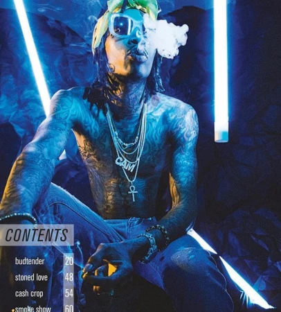 Tattooed adult male seated blowing smoke, wearing sunglasses and jewelry in a blue-lit indoor setting with magazine graphics visible.