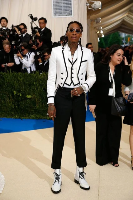 Adult male stands in a white tailcoat and black pants on a white carpet in a room full of photographers at an event.