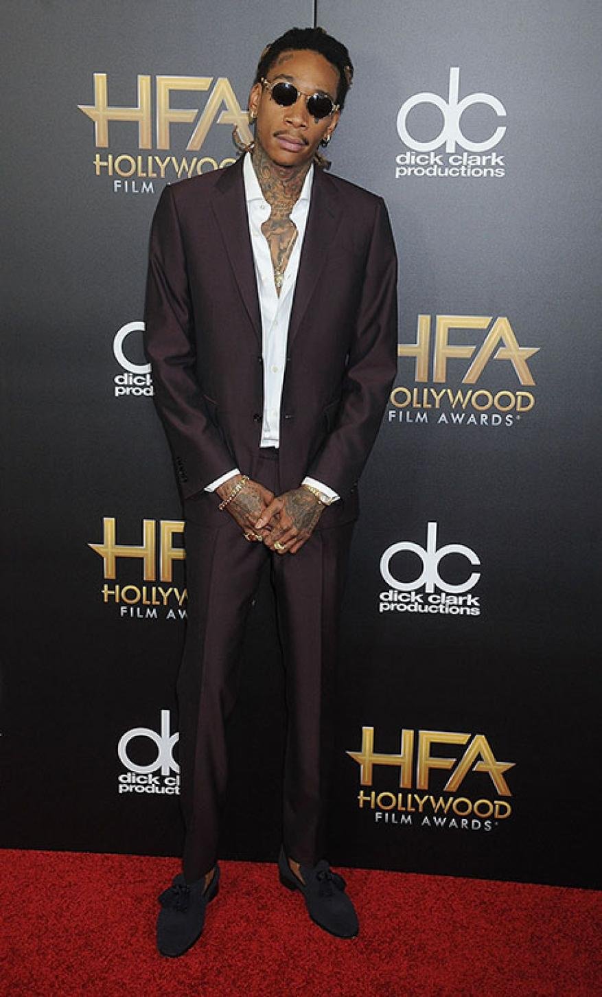 Wiz Khalifa stands on a red carpet at the Hollywood Film Awards in a dark suit, white shirt, sunglasses, and jewelry.