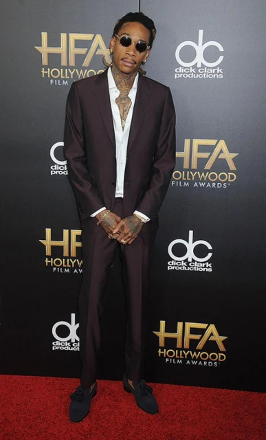 Wiz Khalifa stands on a red carpet at the Hollywood Film Awards in a dark suit, white shirt, sunglasses, and jewelry.