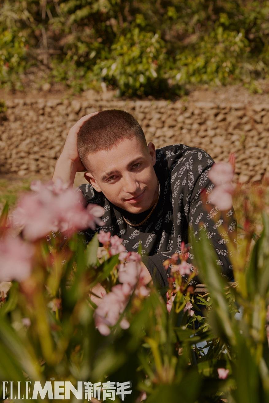 Close-up portrait of a man with short brown hair wearing a patterned dark top, posing among blooming pink flowers outdoors.