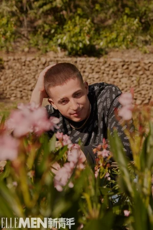 Close-up portrait of a man with short brown hair wearing a patterned dark top, posing among blooming pink flowers outdoors.