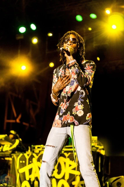 Wiz Khalifa performs on stage at a music concert, wearing sunglasses and a floral shirt with ripped light-wash jeans.