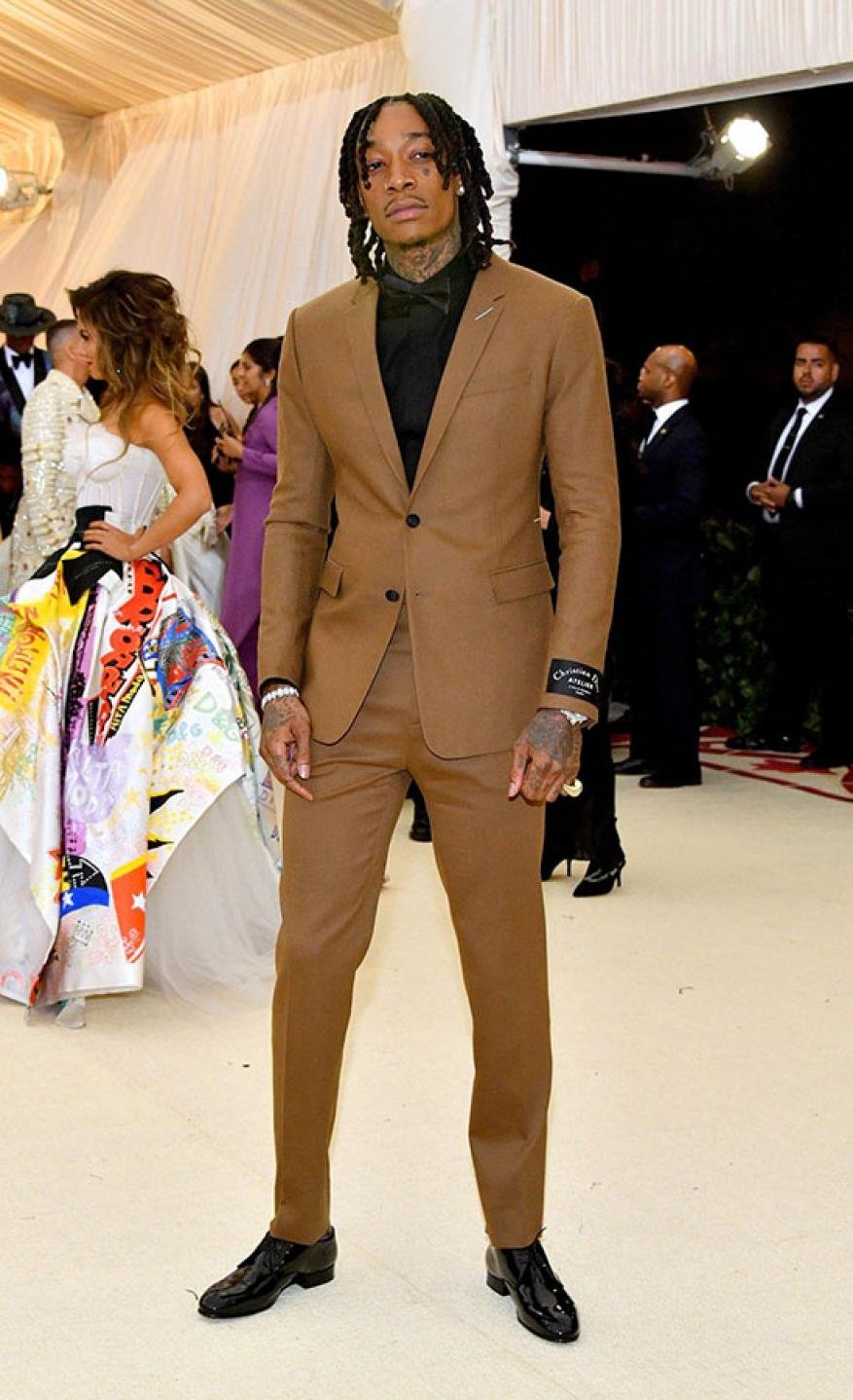 Full-length shot of Wiz Khalifa in a brown suit with a black shirt at an indoor event, surrounded by other attendees and ornate decor.