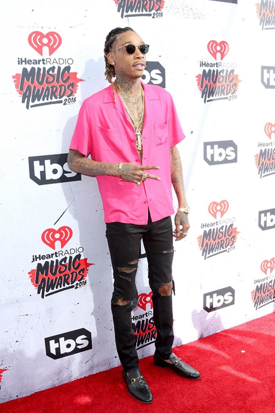 Wiz Khalifa poses in a bright pink shirt and ripped jeans at the iHeartRadio Music Awards, wearing sunglasses and jewelry, on a red carpet.