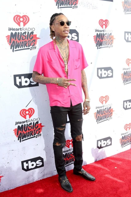 Wiz Khalifa poses in a bright pink shirt and ripped jeans at the iHeartRadio Music Awards, wearing sunglasses and jewelry, on a red carpet.