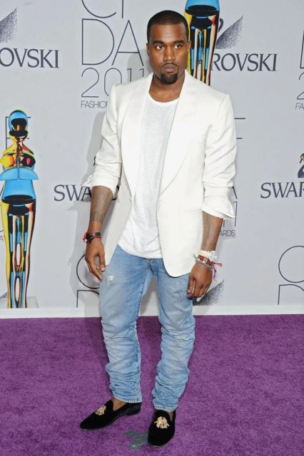 Kanye West poses at an award show, wearing a white blazer, white t-shirt, light blue jeans, and black loafers on a purple carpet.