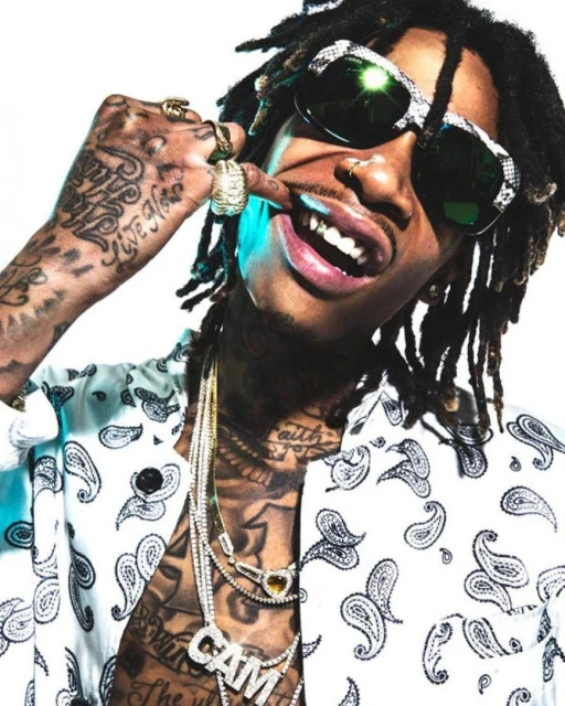 Wiz Khalifa with dreadlocks and sunglasses, wearing a patterned shirt and jewelry, poses against a white background indoors.