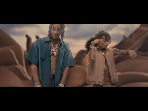 Two men posing in front of a desert landscape with large rock formations, wearing casual clothing and jewelry in a music video scene.