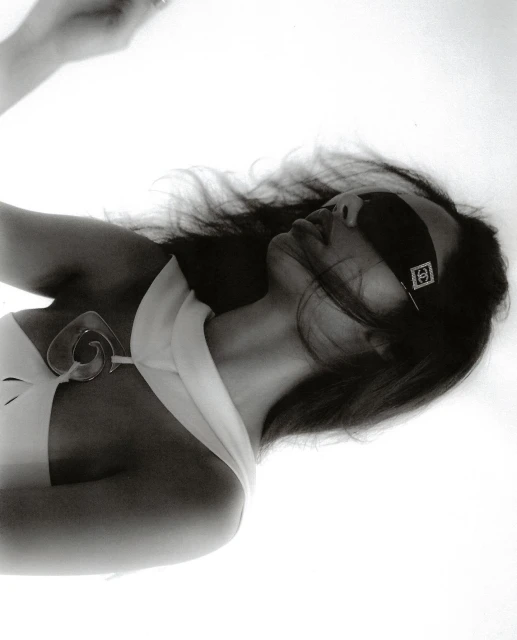 Black and white portrait of a woman with a blindfold and lollipop in an indoor studio environment with bright lighting and minimal props.