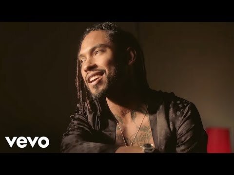 Portrait of an adult male with dreadlocks, fair skin, wearing a satin jacket, smiling in an indoor dark setting with warm lighting.