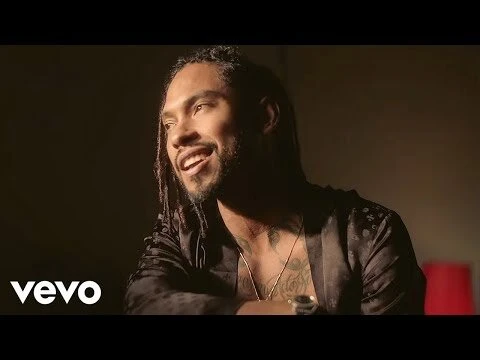 Portrait of an adult male with dreadlocks, fair skin, wearing a satin jacket, smiling in an indoor dark setting with warm lighting.