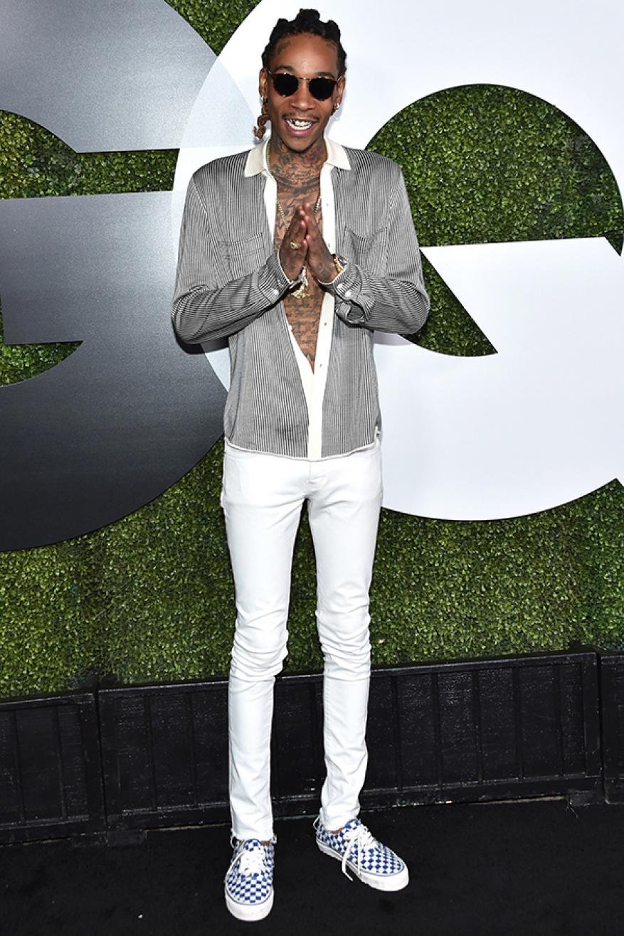 Full-length shot of Wiz Khalifa at GQ event, wearing sunglasses, button-down shirt, white pants, and checkered shoes, against logo backdrop.
