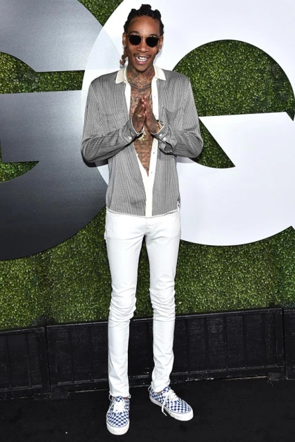 Full-length shot of Wiz Khalifa at GQ event, wearing sunglasses, button-down shirt, white pants, and checkered shoes, against logo backdrop.