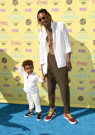 Wiz Khalifa and a young child pose on a blue carpet in front of a yellow Teen Choice Awards backdrop with sponsor logos.