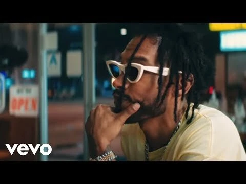 Adult male with dreadlocks and sunglasses, wearing a yellow shirt indoors, posing near an open sign and a window at night.
