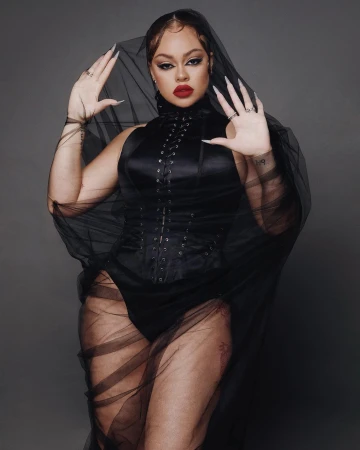 Woman wearing a black lace veil and black lace bodysuit with corset styling, posed against a dark grey background.