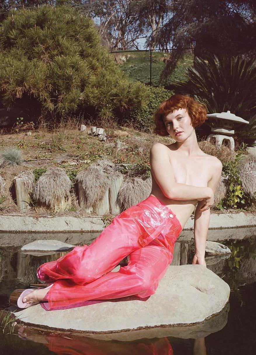 A red-haired adult woman poses topless on a rock, wearing transparent pink pants, set within a serene Japanese garden with pond and greenery.