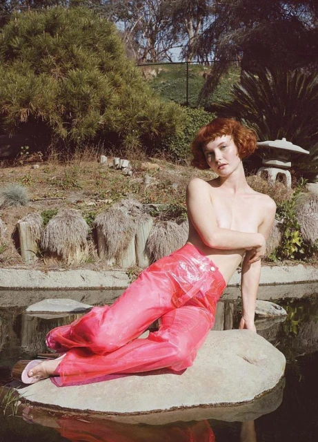 A red-haired adult woman poses topless on a rock, wearing transparent pink pants, set within a serene Japanese garden with pond and greenery.