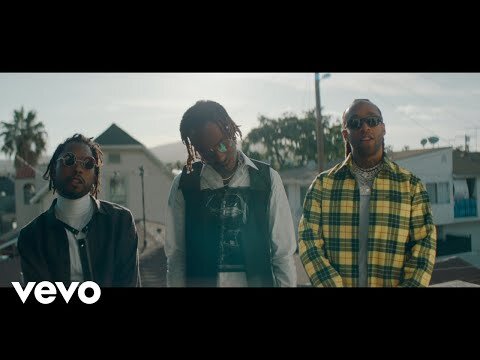 Three adult men stand outdoors near houses and palm trees wearing fashionable clothing and sunglasses in a music video scene.