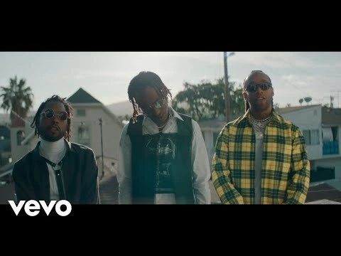 Three adult men stand outdoors near houses and palm trees wearing fashionable clothing and sunglasses in a music video scene.