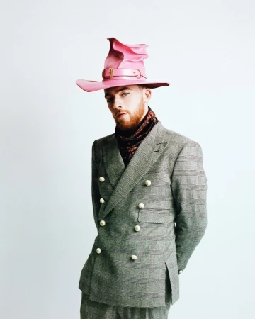 Angus Cloud wearing a pink leather witch hat and grey double-breasted plaid suit styled by Lauren Matos against white