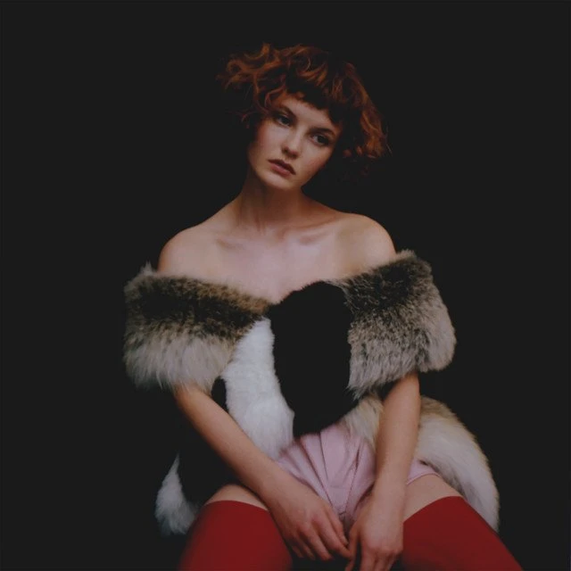 A woman with red hair, wearing a fur shawl, pink shorts, and red stockings, poses in front of a dark background in a portrait.