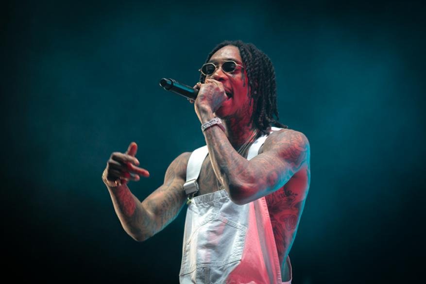 A photo of Wiz Khalifa performing on stage, wearing overalls and sunglasses, holding a microphone, and gesturing with his left hand.