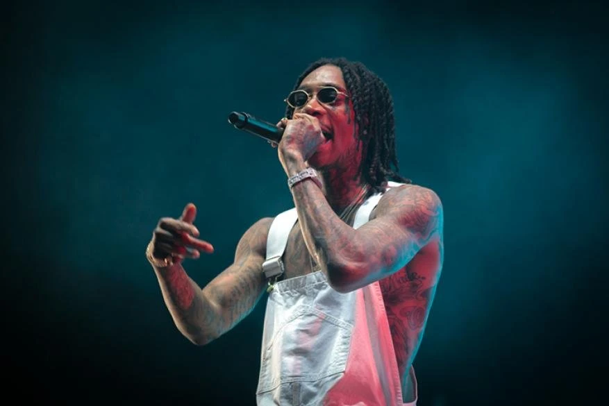A photo of Wiz Khalifa performing on stage, wearing overalls and sunglasses, holding a microphone, and gesturing with his left hand.