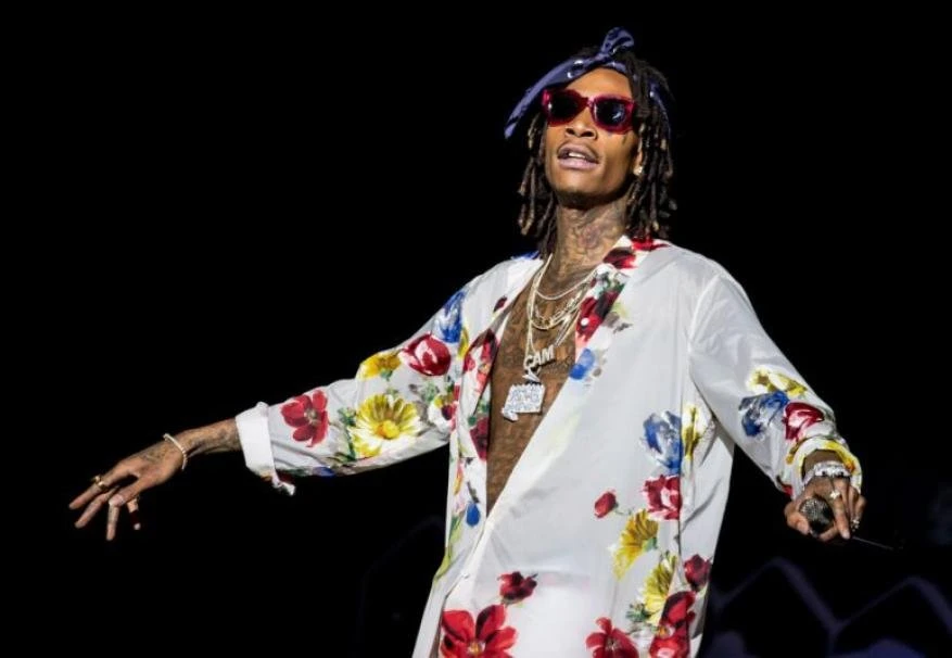 Adult male entertainer performing on stage wearing floral shirt, headscarf, sunglasses, and multiple chains in a dimly lit indoor environment.