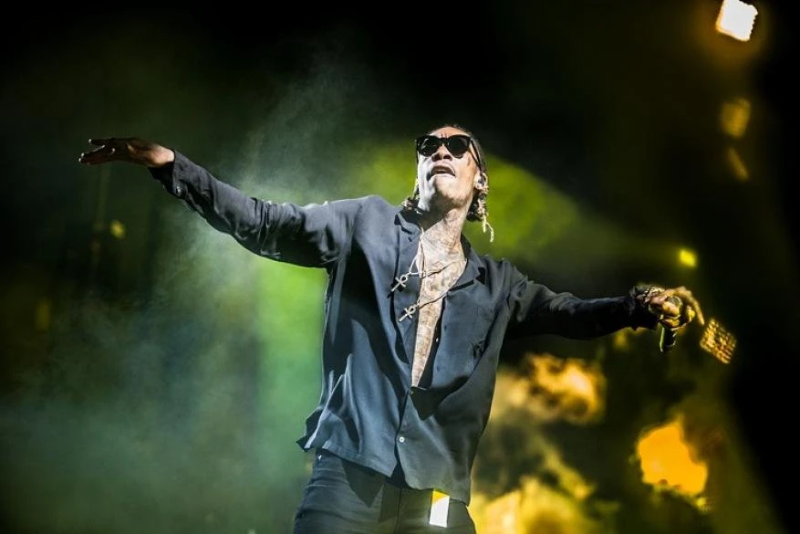 Adult male wearing sunglasses and a black shirt performing on a stage with arms outstretched, bathed in bright yellow stage lighting.