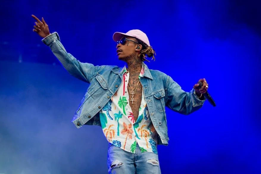 Wiz Khalifa performing on stage, wearing a pink hat, sunglasses, denim jacket, and tropical-print shirt, against a solid blue background.