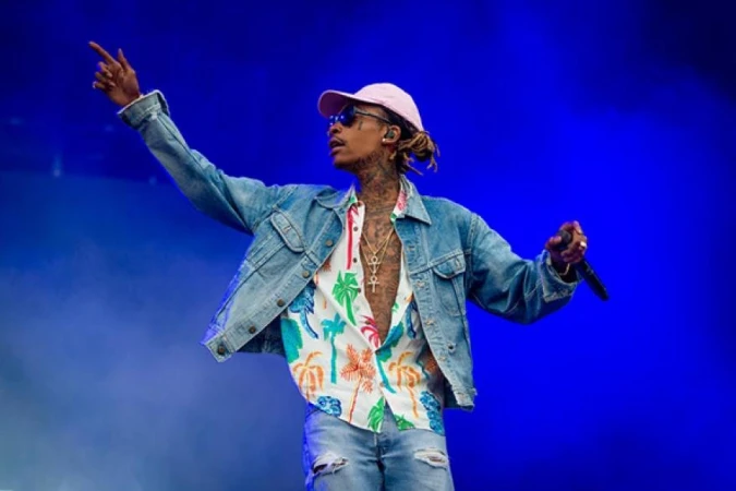 Wiz Khalifa performing on stage, wearing a pink hat, sunglasses, denim jacket, and tropical-print shirt, against a solid blue background.