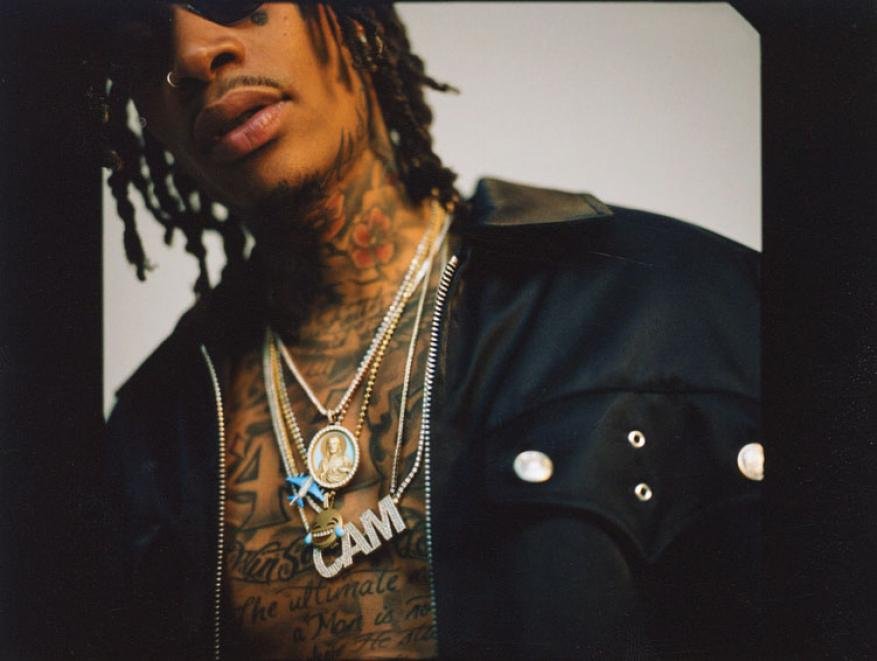 Portrait of a man with dreadlocks, tattoos, and multiple chains. He is wearing a black jacket against a light background.