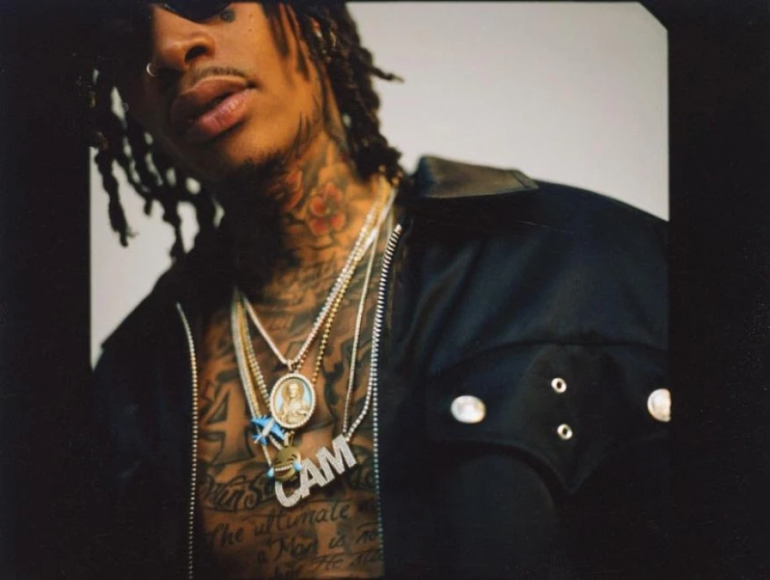 Portrait of a man with dreadlocks, tattoos, and multiple chains. He is wearing a black jacket against a light background.