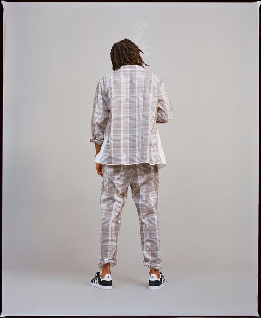 Back view of a person with dreadlocks wearing a white and brown plaid suit and Adidas sneakers standing in a neutral photo studio.