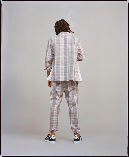 Back view of a person with dreadlocks wearing a white and brown plaid suit and Adidas sneakers standing in a neutral photo studio.