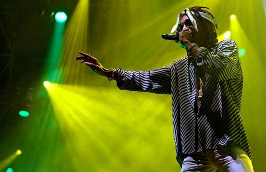 A man wearing sunglasses and a patterned striped shirt holding a microphone on stage under bright yellow stage lights.