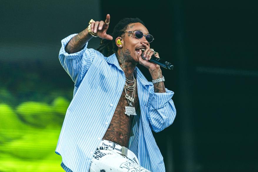 Wiz Khalifa performing live on stage with a microphone, wearing sunglasses, an open blue striped shirt, and white patterned pants.