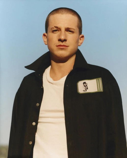 Portrait of an adult male with a buzzcut, wearing a dark jacket over a white t-shirt, posing against a clear blue sky backdrop.