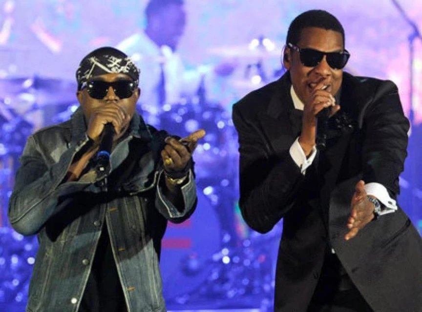 Two adult males, wearing sunglasses, perform on stage. One in a suit, the other in a denim jacket, both holding microphones and gesturing.
